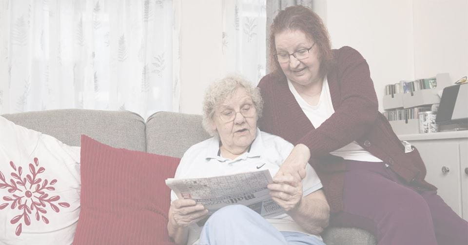 Ældre kvinder kigger i avis, mens en anden kvinde sidder ved siden af hende og peger i samme avis